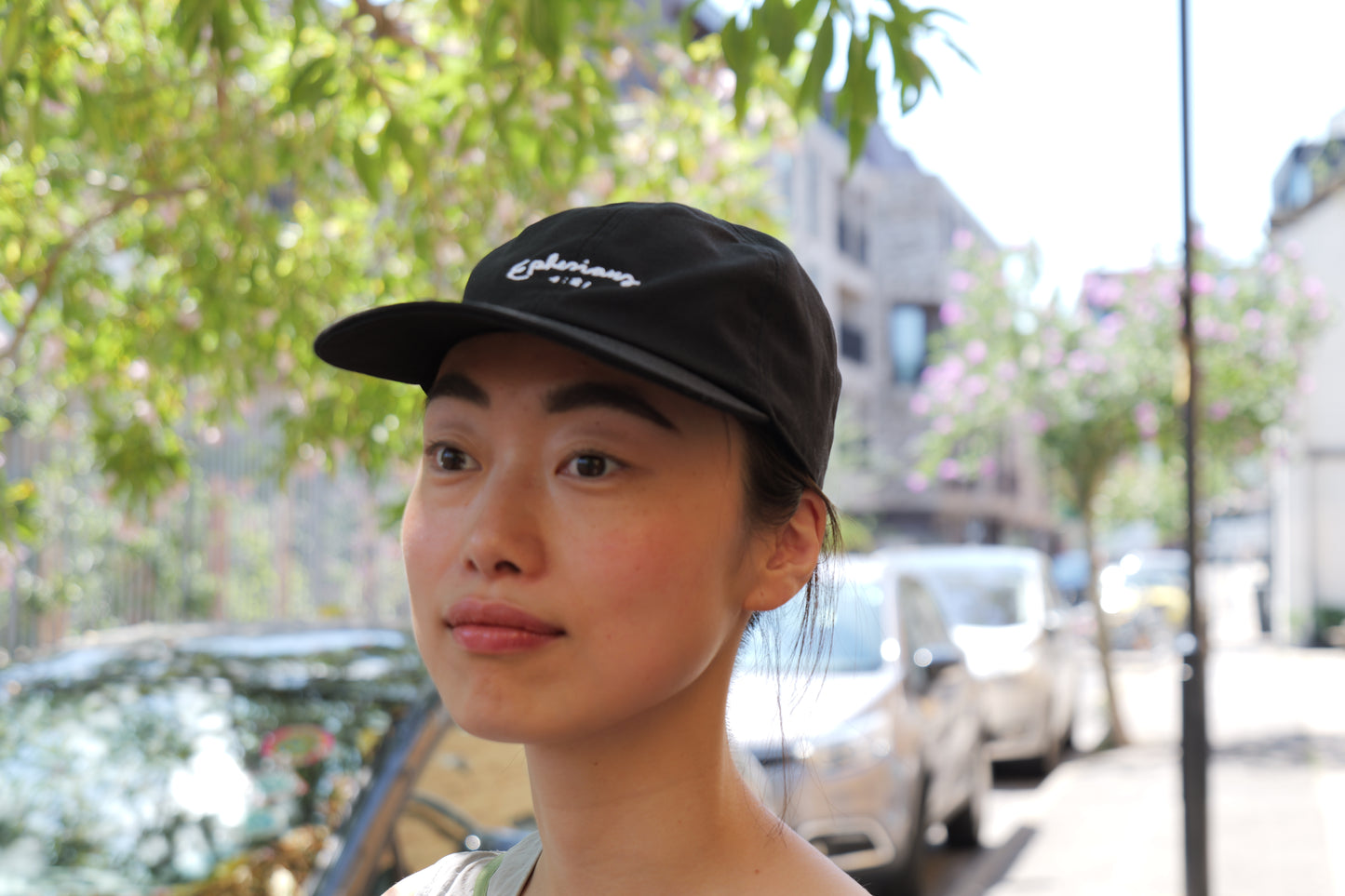Person wearing a black cap with text, standing outdoors with trees and buildings in the background