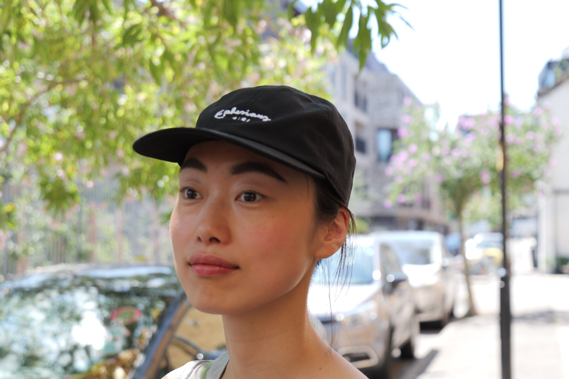Person wearing a black cap with text, standing outdoors with trees and buildings in the background