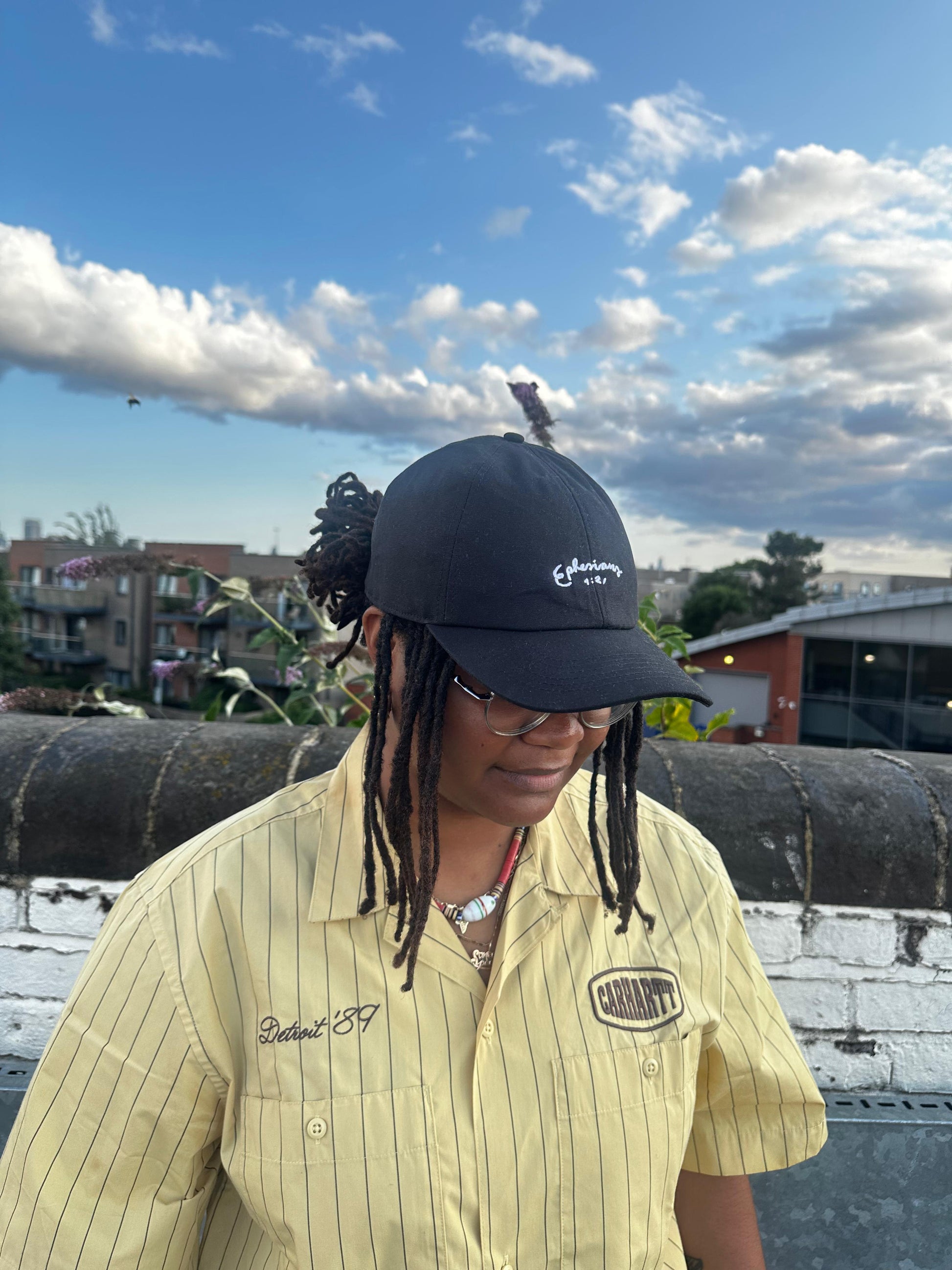 Person wearing a black cap and yellow striped shirt on a rooftop with a blue sky and clouds in the background.